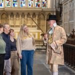 A man in Regency costume points a cane while a group of 3 women and 1 man listen to him talking in a church.