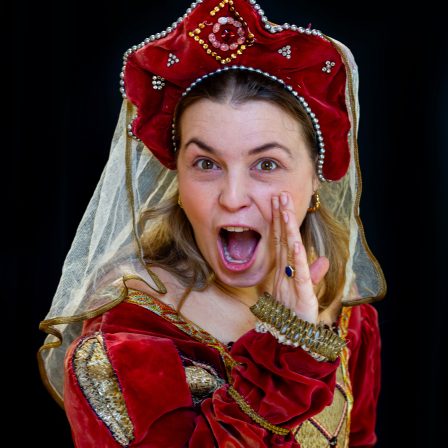 A woman in a red medieval dress, hat and veil looks like she is whispering an exciting secret