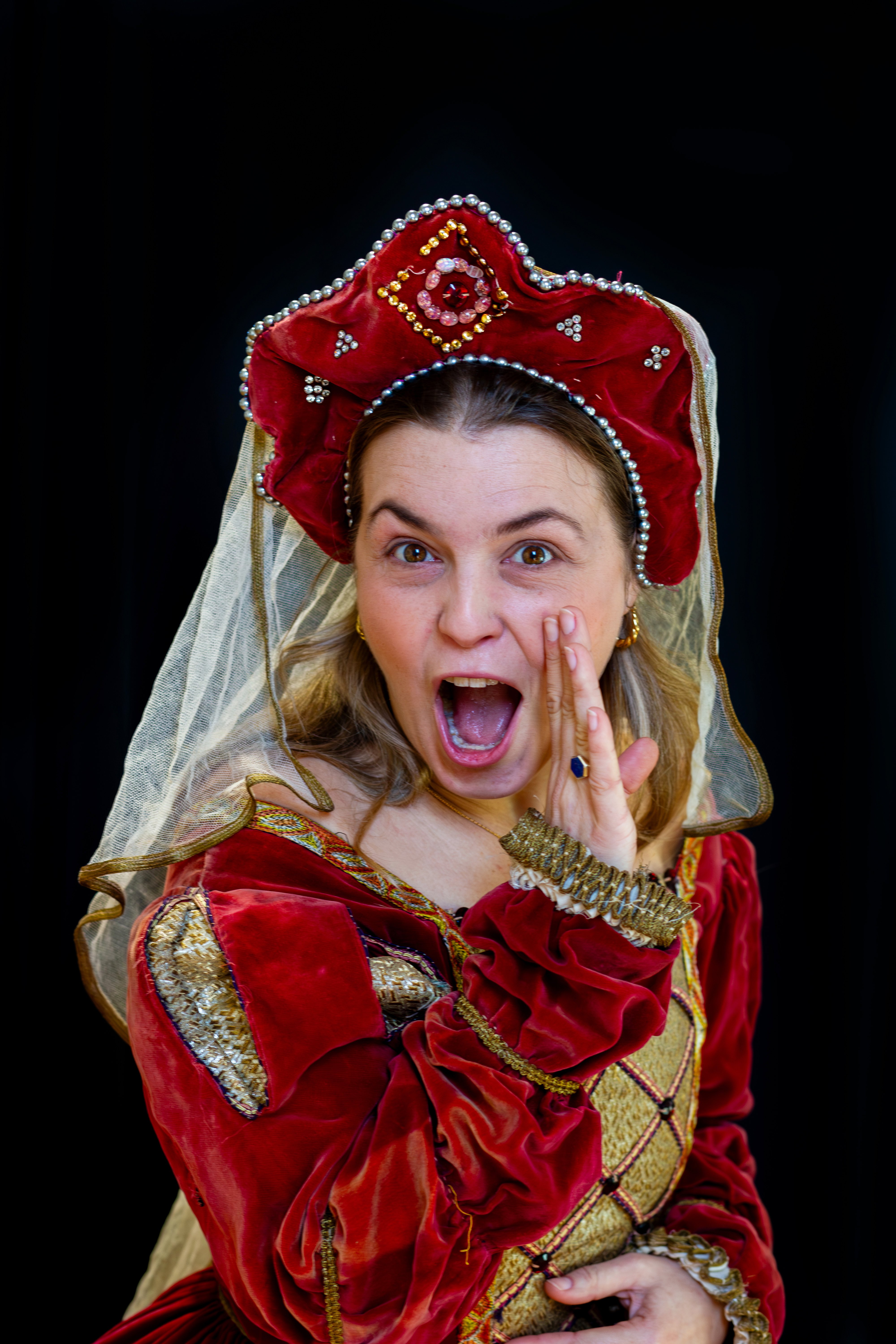 A woman in a red medieval dress, hat and veil looks like she is whispering an exciting secret